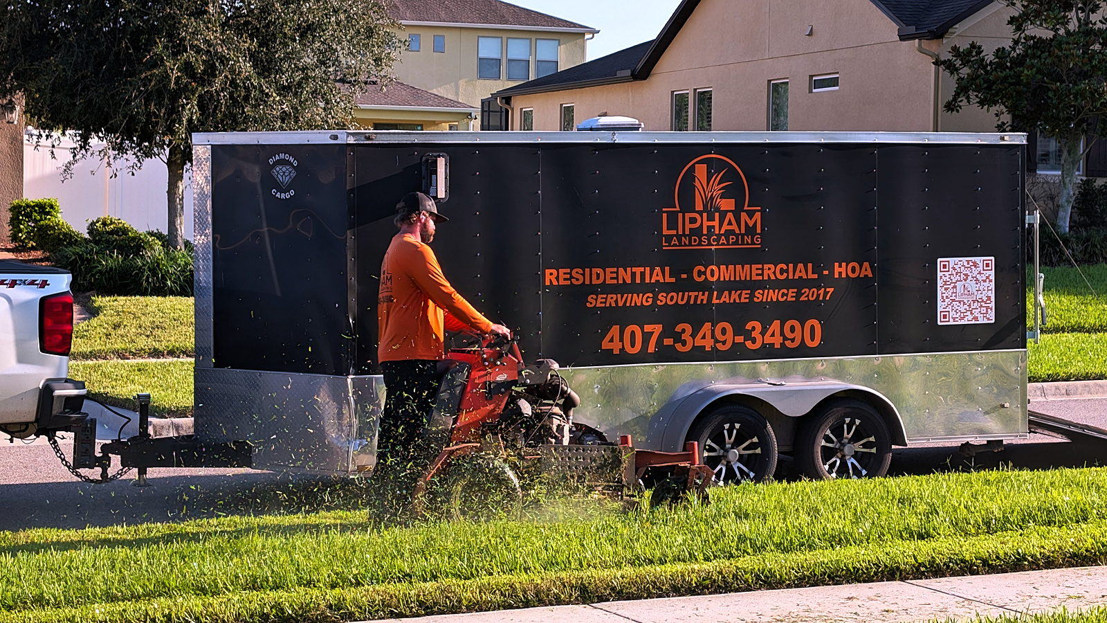 A Lipham Landscaping crew member mowing beside the company's branded trailer, clippings in flight